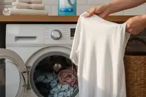 Hands hold a wet white T-shirt in front of an open front-loading washing machine filled with clothes, with folded towels on a shelf above.